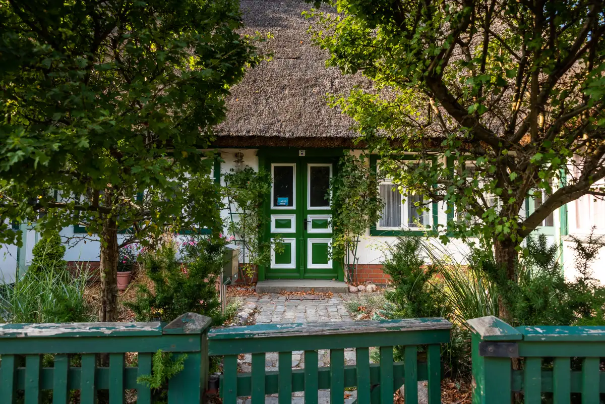 Haus mit Reetdach und grüner Doppeltür zwischen Bäumen, davor ein grüner Holzzaun
