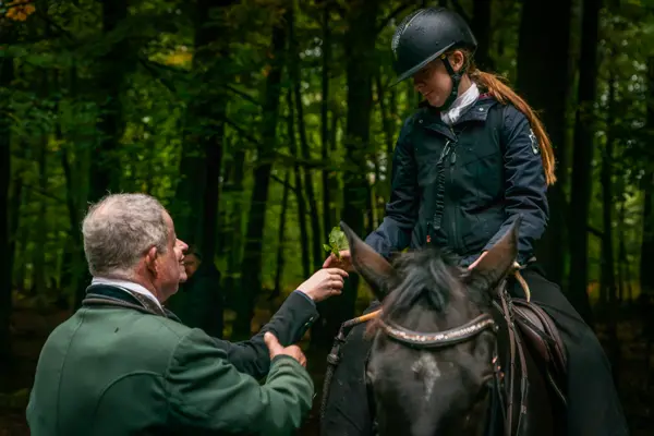 Ein Mann im grünen Rock reicht einer Reiterin im Wald einen Zweig