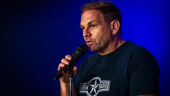 Quizmaster Dominik Bartels spricht ins Mikrofon, hinter ihm blaues Bühnenlicht