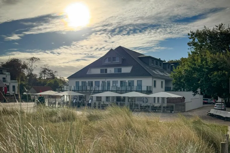 Strandrestaurant Achterdeck in Graal-Müritz: weißes Haus mit dunklem Dach und Sonnenschirm-Terrasse hinter Dünengras im Gegenlicht